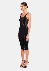 Schwarzes, figurbetontes Kleid mit transparenten Einsätzen, Rundhalsausschnitt und knielangem Saum. Texturkontraste zwischen festem Material und Mesh. Hohe Sandalen.