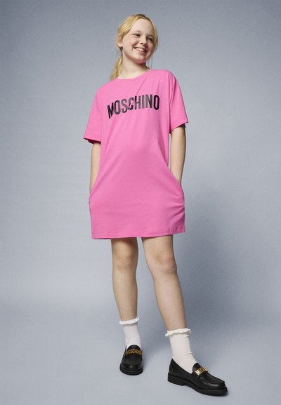 Rožinė medvilninė t-shirt suknelė trumpomis rankovėmis, su juodu "Moschino" logotipu. Su ja derinamos juodos loafer batai ir baltos pūkuotos kojinės.