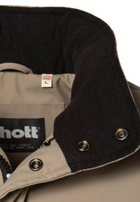 Giacca beige con colletto foderato in fleece nero, dotata di bottoni a pressione dorati e di un'etichetta che mostra la taglia "L" e il design della bandiera americana.
