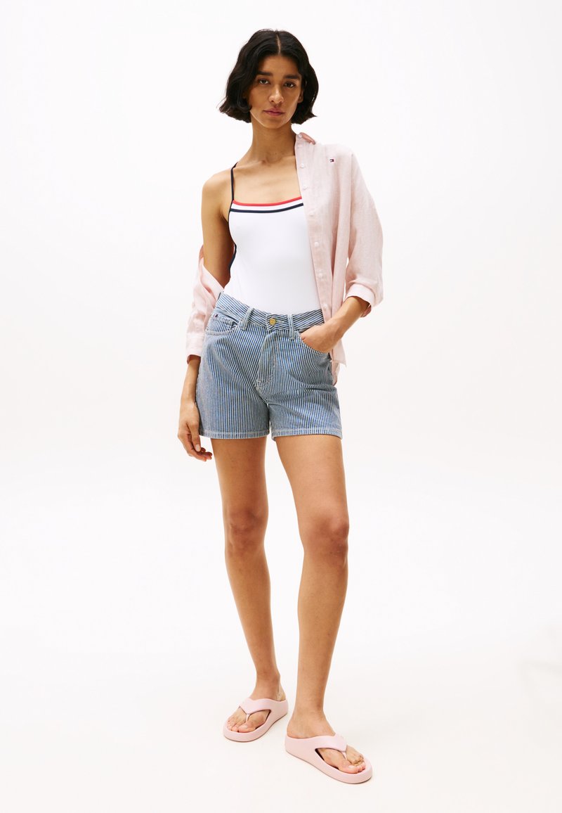 Costum de baie alb cu dungi roșii și bleumarin, cămașă deschisă roz, pantaloni scurți rayati albastre și slapi roz deschis. Vestimentație simplă, casual pentru plajă.