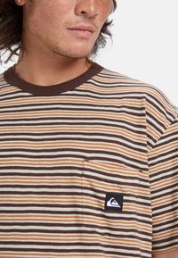 Camiseta de rayas en colores marrón, blanco y naranja; de manga corta; con cuello redondo de canalé marrón; presenta un bolsillo en el pecho y un logo.