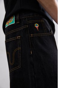 Black denim jeans with yellow stitching, featuring a colorful ice cream cone embroidery on the back pocket and a patch on the waistband.