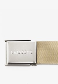 Béžový pletený opasok so lesklou striebornou kovovou prackou s vygravírovaným názvom značky "LACOSTE." Textúrovaná tkanina pre odolnosť.