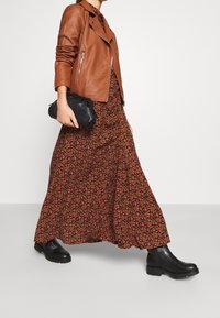 Giubbotto in pelle marrone sopra un abito maxi nero a fiori. Stivali neri alla caviglia e una piccola borsetta nera completano l'outfit.