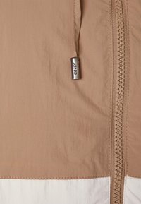 Tan jacket with a smooth texture, featuring a prominent zipper, silver hardware, and a contrasting white lower section.