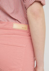 Pantalones de mezclilla rosa con una etiqueta de cuero beige que dice "BOSS" en la parte de atrás. Presentan bolsillos cosidos y una textura suave. Camisa a rayas parcialmente visible.