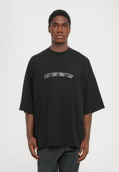 Young man wearing an oversized black t-shirt with white "032C" logo on the chest, standing against a plain light background.