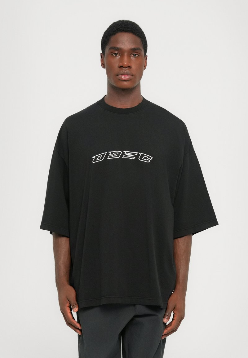 Jonge man in een overgrote zwarte t-shirt met een witte "032C" logo op de borst, staande tegen een eenvoudige lichte achtergrond.