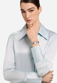 Bluză din satin de culoare albastru deschis, cu guler și cu nasturi. O brățară argintie cu un cadran rectangular și link-uri de lanț împodobește încheietura.