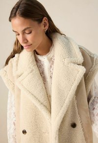 Femme regardant vers le bas, portant un gilet en shearling crème sur une blouse blanche en dentelle boutonnée, contre un fond uni.