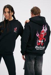 To personer iført sorte hættetrøjer med en rød tegneserie-djævel og teksten "Slip din indre djævel løs", den ene viser forsiden, den anden viser bagsiden.