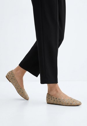Persona con zapatos planos beige con detalles de ojales metálicos y pantalones negros, caminando sobre un suelo blanco.