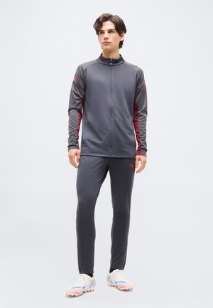 Jeune homme debout portant une veste zippée gris foncé et un pantalon assorti avec des détails rouges, accompagné de chaussures de sport blanches sur un fond clair.