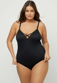 Femme portant un maillot de bain une pièce noir avec des fines bretelles et des détails en perles dorées le long du décolleté en V profond, posant devant un fond uni.