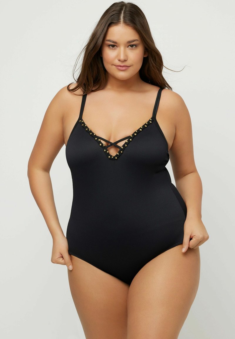 Femme portant un maillot de bain une pièce noir avec des fines bretelles et des détails en perles dorées le long du décolleté en V profond, posant devant un fond uni.