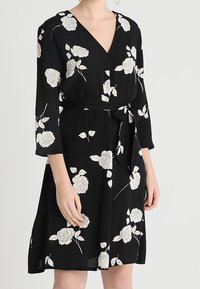 Vestido negro con escote en V, mangas tres cuartos y cintura con lazo. Presenta patrones florales blancos en toda su superficie. Textura suave.