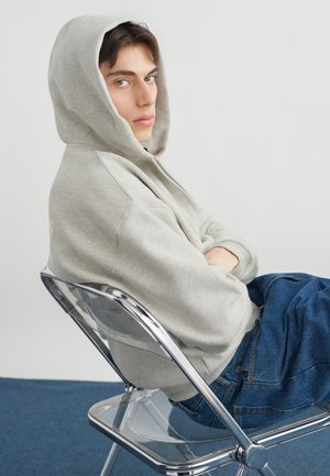 Noor hall halli ja sinistes teksades, istub külili läbipaistval toolil, käed ristatud, vaatab kaamerasse.