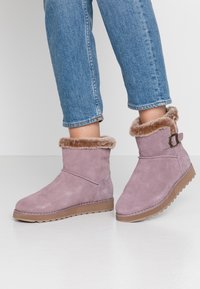 Fialové semišové kotníkové boty s koženým lemem, s postranním sponou a texturovanou gumovou podrážkou pro lepší přilnavost. Nosené s zkrácenými modrými džíny.