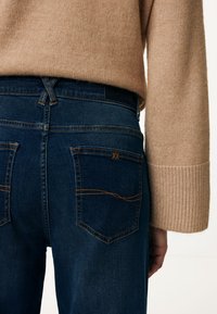 Mörkblå denimjeans med orange sömmar, med två bakfickor och en synlig midja ovanför en beige tröja.
