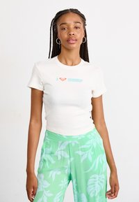 T-shirt crème à manches courtes avec le texte "J ❤️ surf" en bleu clair et un logo de cœur rouge. Associé à un pantalon large à motif floral vert.