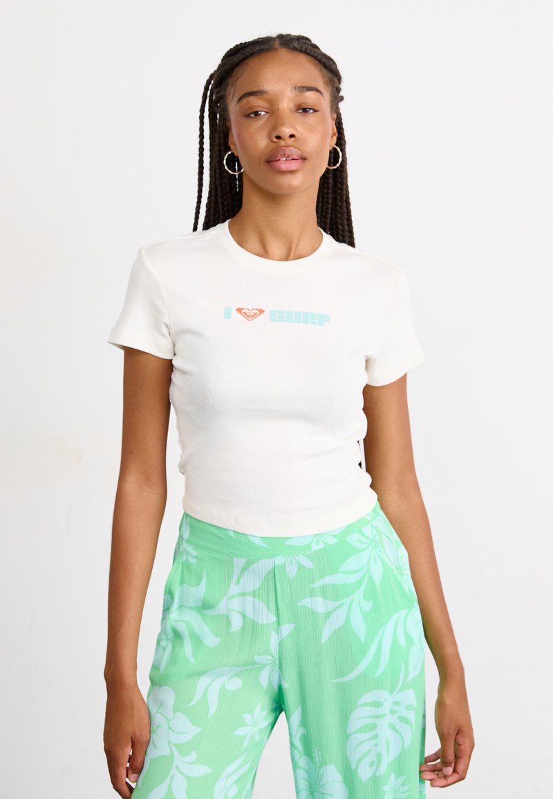 T-shirt crème à manches courtes avec le texte "J ❤️ surf" en bleu clair et un logo de cœur rouge. Associé à un pantalon large à motif floral vert.