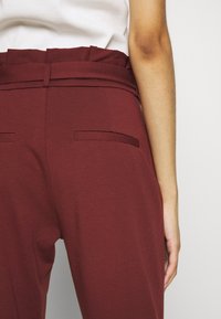 Pantalon bordeaux taille haute avec une texture lisse, présentant deux poches arrière et un détail de ceinture, vu de dos.