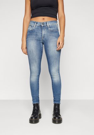 Jeans ajustados de denim azul claro con un patrón desvaído. Cuenta con cinco bolsillos y cierre de botón. Se llevan con botas de combate negras.