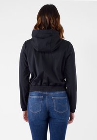 Giacca nera leggera con cappuccio, vita elastica e polsini aderenti, abbinata a jeans di denim blu con tasche posteriori.