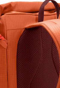 Mochila de tela naranja con una superficie texturizada, que cuenta con un forro de malla acolchada en color burdeos y una correa ajustable para mayor comodidad.