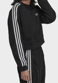 Svart kort sweatshirt med långa ärmar, med tre vita vertikala ränder på varje axel och en broderad logotyp på bröstet.