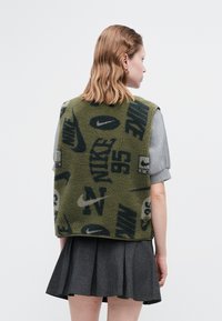 Prsluk od flisa u maslinasto zelenoj boji s crnim Nike logotipima i uzorcima. Ima kvadratni kroj i nosi se preko sive majice s kratkim rukavima.