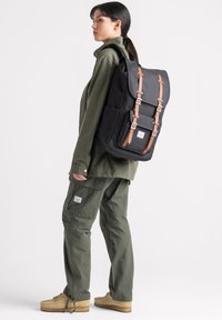 Schwarzer Canvas-Rucksack mit braunen Lederträgern und -schnallen, ausgestattet mit einer Fronttasche und rechteckiger Form, einseitig über einer Schulter getragen.