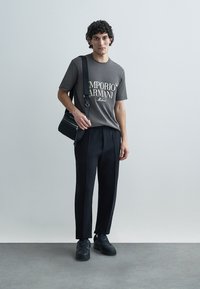 Gray t-shirt with white text logo, black pleated trousers, and black sneakers. Model holds a black crossbody bag. Simple background.