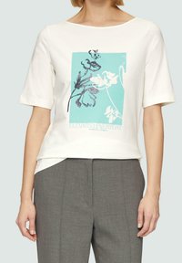 Vit kortärmad t-shirt med en turkos blommig grafik med glittrande detaljer och texten "ELEVATED EVERYDAY" längst ner.