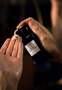 Baardvochtinbrengende crème in een donkerblauwe fles met een pompdeksel. Witte crème uitgestort op een palm, met een zilveren label en zwarte tekst.