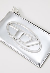 Silver metallic pouch with a smooth texture, featuring a prominent silver logo detail. Edges are finished with white stitching.