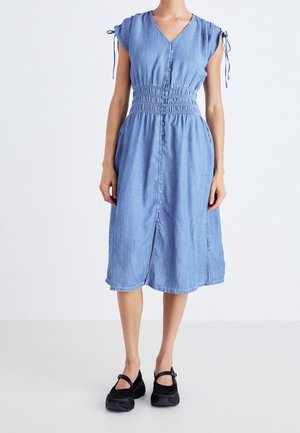Femme portant une robe midi boutonnée bleu clair avec une taille froncée et des manches courtes, associée à des chaussures Mary Jane noires.