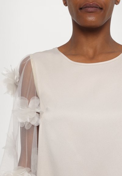 Light beige blouse featuring sheer, textured sleeves adorned with layered, white floral appliqués and a rounded neckline.