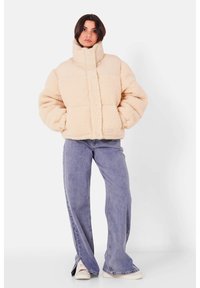 Beige, fluffy pufferjakke med høj krave og trykknaplukning. Har en struktureret krop og en glat overdel. Bæres med blå brede jeans.