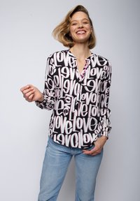 Blouse à manches longues noire avec un motif "love" blanc et rose ; avec une fermeture à boutons sur le devant et une coupe décontractée. Associée à un jean bleu clair.