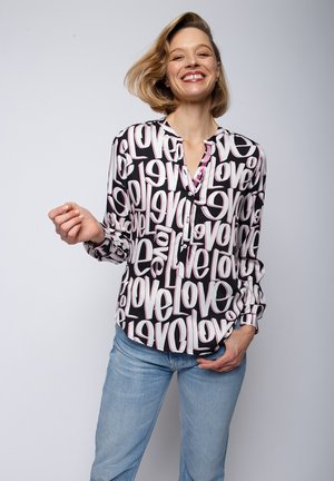 Blouse à manches longues noire avec un motif "love" blanc et rose ; avec une fermeture à boutons sur le devant et une coupe décontractée. Associée à un jean bleu clair.