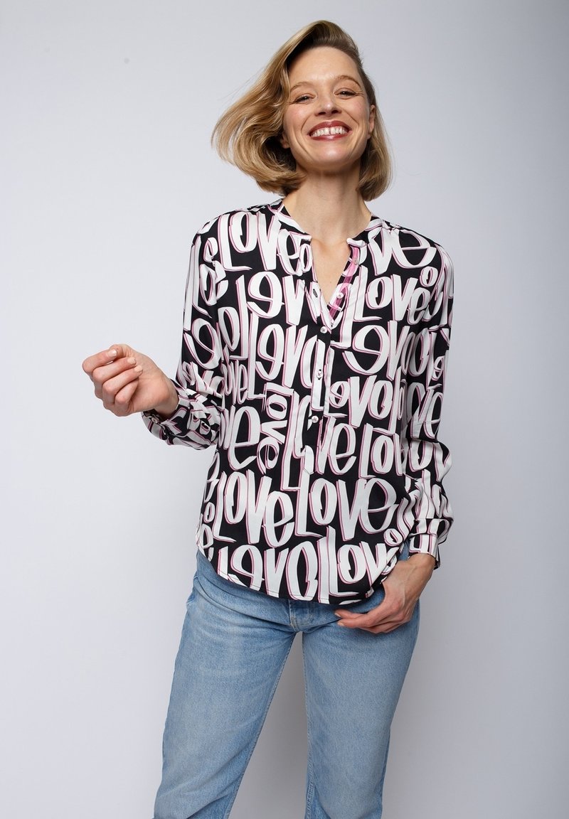 Blouse à manches longues noire avec un motif "love" blanc et rose ; avec une fermeture à boutons sur le devant et une coupe décontractée. Associée à un jean bleu clair.