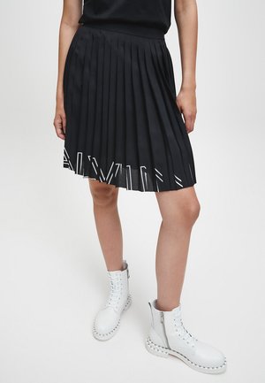 Personne portant une jupe plissée noire avec un détail de texte blanc près de l'ourlet et des bottines blanches à lacets avec des fermetures éclair latérales.