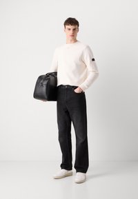 Witte lange mouwen trui met V-hals, zwarte spijkerbroek en witte sneakers. Model houdt een zwarte leren tas vast met een rits en ronde vorm.