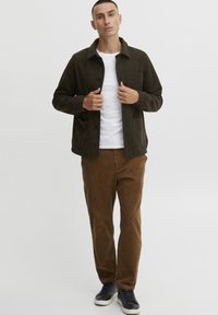 Chaqueta texturizada verde oscuro con cierre de botón en el frente y bolsillo en el pecho, llevada sobre una camiseta blanca y combinada con pantalones de pana marrones y zapatillas.