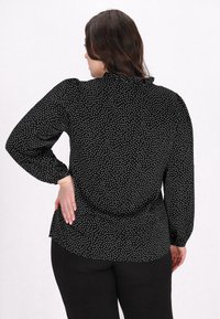 Schwarze Bluse mit weißen Punkten, langen Ärmeln mit elastischen Bündchen, abgerundetem Saum und hohem Kragen. Das Material scheint leicht und strukturiert zu sein.