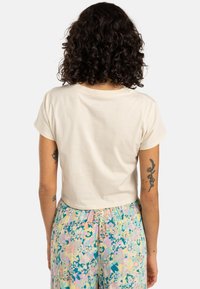 Crème kleurig, cropped t-shirt met korte mouwen, gladde katoenstructuur, gepositioneerd tegen een neutrale achtergrond, gedragen met gebloemde, gemusterde broek.