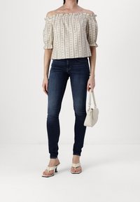 Haut à motifs floraux avec épaules dénudées et manches bouffantes, associé à un jean skinny foncé et des sandales blanches à talons. Tient un petit sac à main beige.