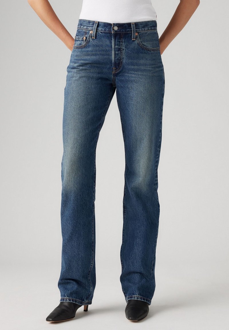 Personne portant un jean bleu droit, un haut blanc et des chaussures noires à talons, debout les mains dans les poches.