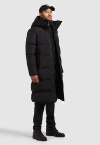 Sort pufferjakke med lang design, der har hætte og lynlås. Båret over en sort top, parret med mørke jeans og sorte støvler.